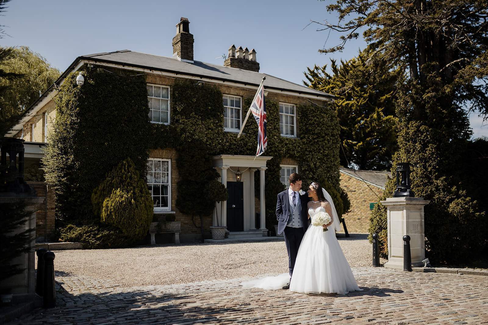 A classic portrait of a bride and groom walking together and chatting naturally in front of the historic Friern Manor house.