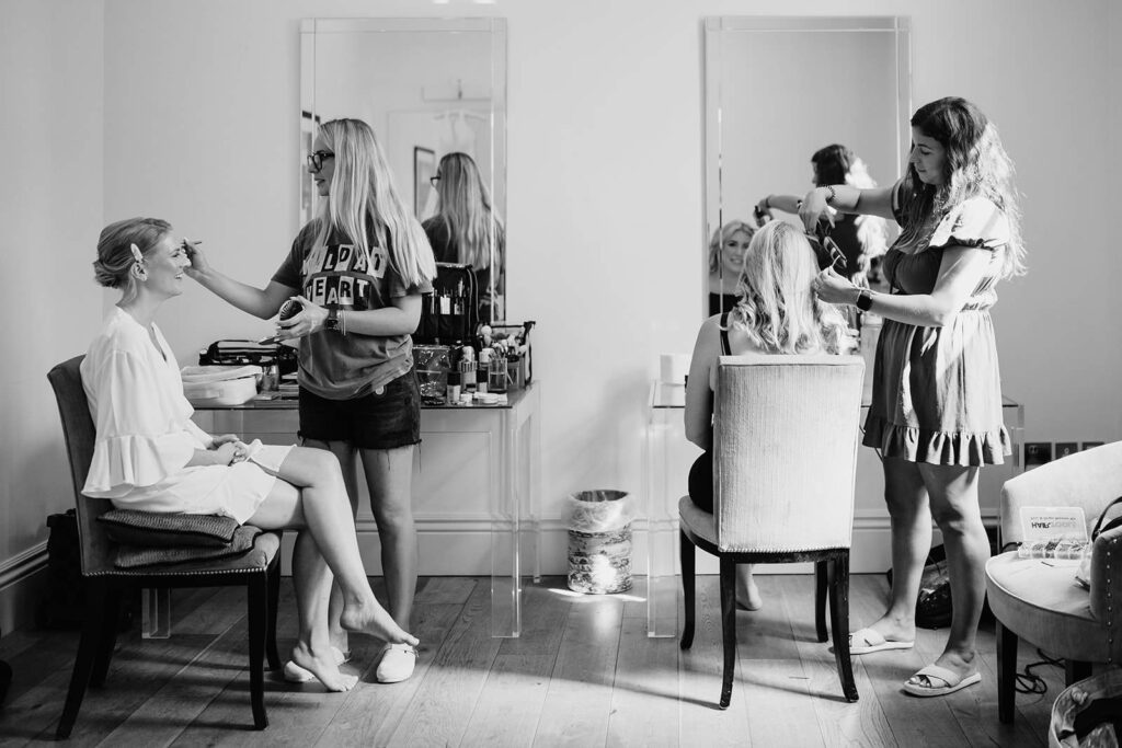 A black and white photograph of a bride and her bridesmaid sitting in chairs while having their makeup applied in the bridal preparation room at Braxted Park.