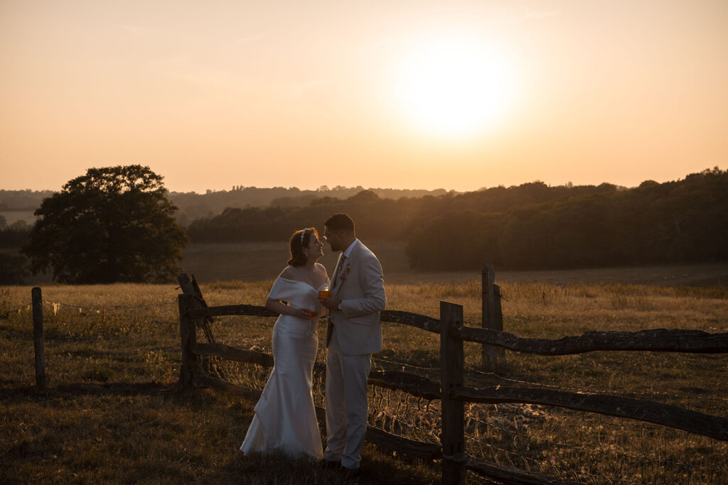 A romantic 2026 wedding photograph of Harriet and Joe in a sunset portrait at Gaynes Park, Essex; bride and groom silhouetted against the golden hour light in the Epping meadows.