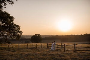 A wide-angle 2026 wedding photograph of Harriet and Joe standing in the vast golden hour meadows of Gaynes Park, Essex, captured in a cinematic, environmental style.