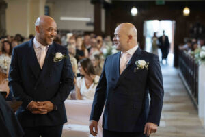 A 2026 black and white wedding photograph of a mature groom (Darren) and his best man sharing a moment of quiet confidence inside the historic St Anne’s Church, Limehouse.