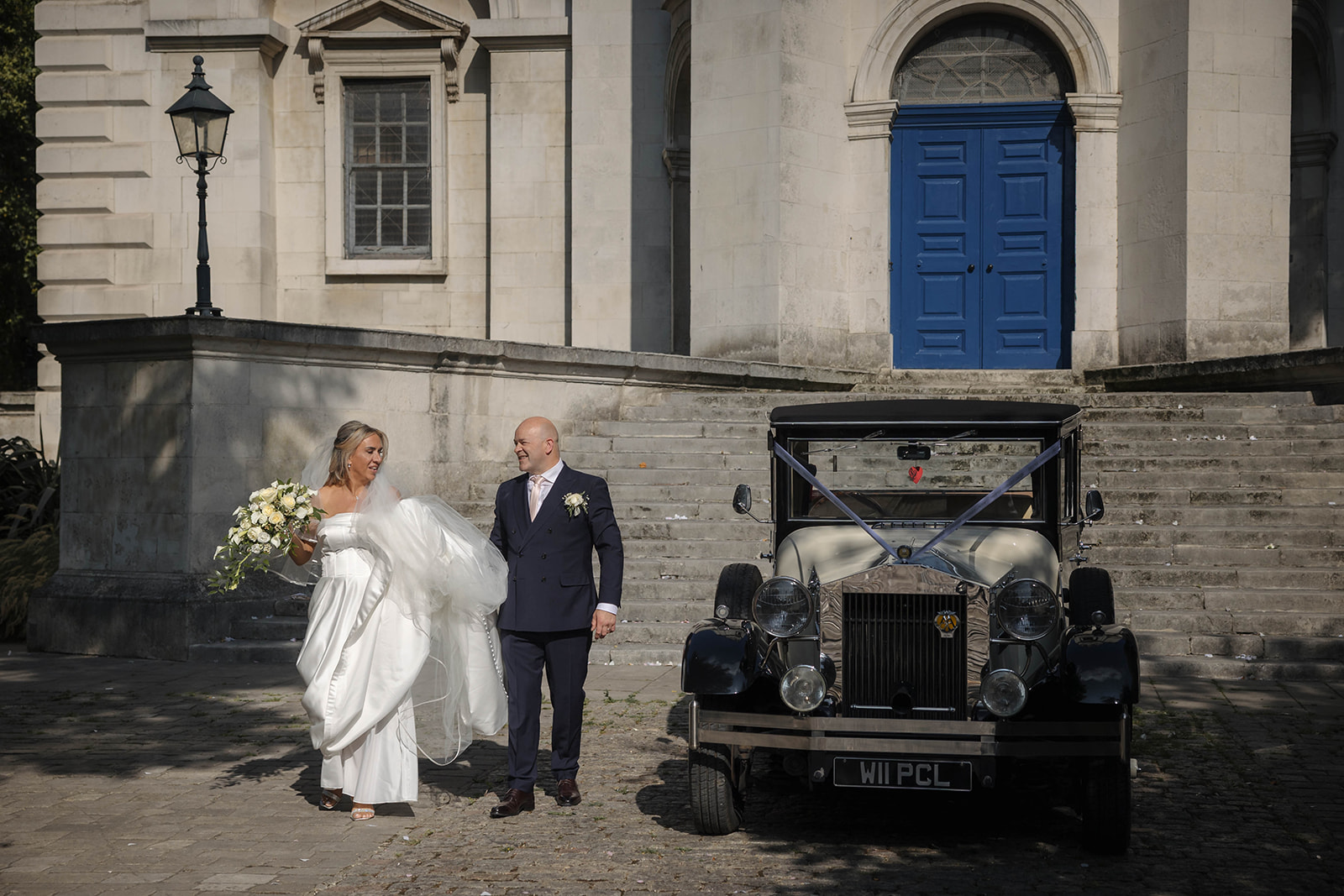 A 2026 London wedding photograph of Melissa and Darren walking next to their vintage wedding car outside the historic St Anne’s Church, Limehouse.