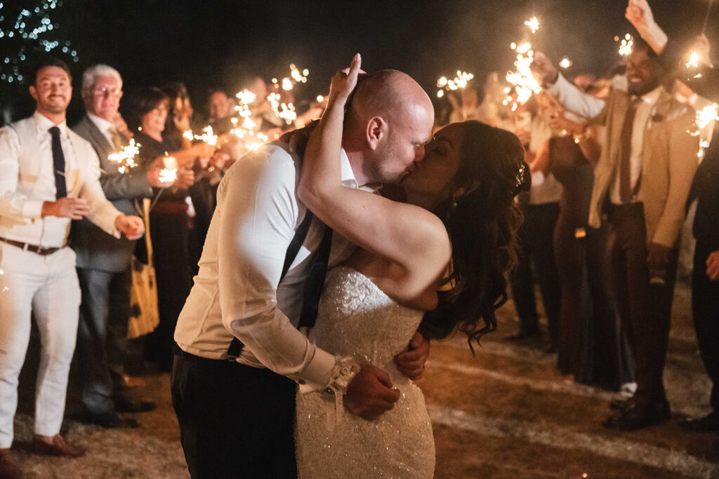 Wedding photography Creeksea Place; cinematic night shot of a bride and groom kissing surrounded by guests with sparklers at an Essex wedding venue.